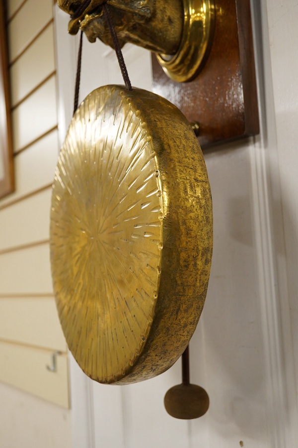 An early 20th century brass wall hanging gong, the holder in the form of a big cat, mounted on an oak plinth, the gong is supported on a bone clenched between its teeth, with the beater on a separate hook behind, 55cm to